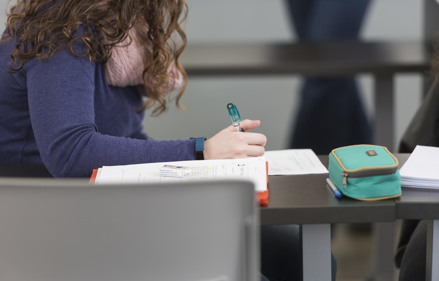 A cropped image of someone holding a pencil in their hand while sitting at a desk working on homework.