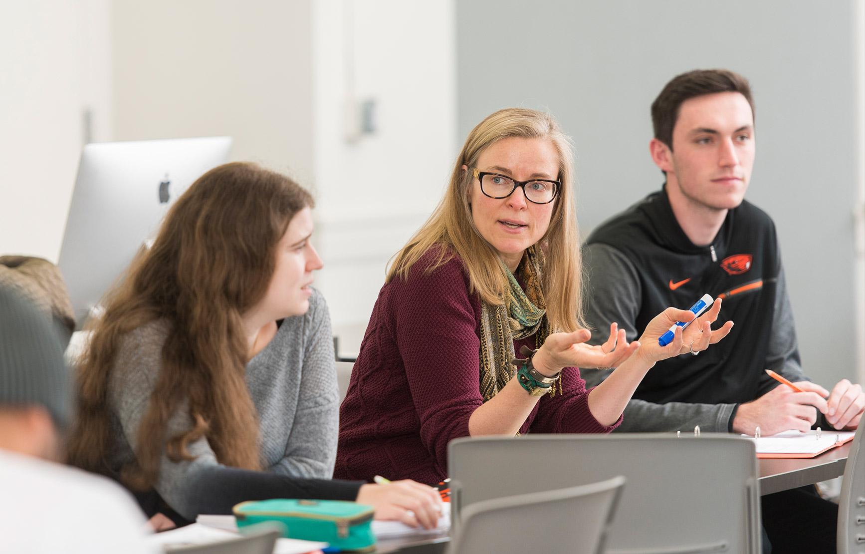 A woman professor sits between two students in a computer lab classroom, explaining concepts